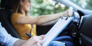 Close up view of driving instructor holding checklist while in background female student steering and driving car.